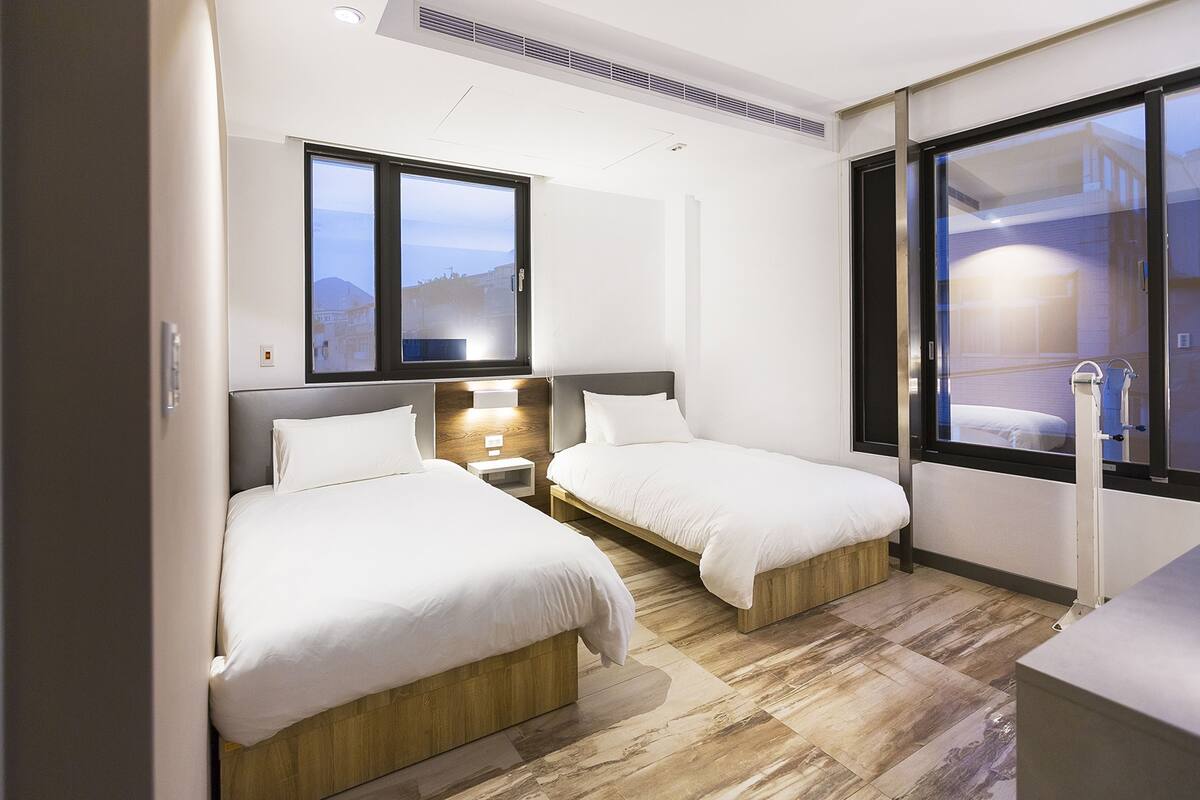 A hotel room with two beds, a small bedside table, a large window, and a view of the outside.