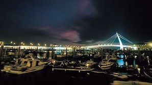 Un port de plaisance illuminé la nuit, avec des bateaux amarrés, un pont décoré de lumières et une lune dans le ciel.