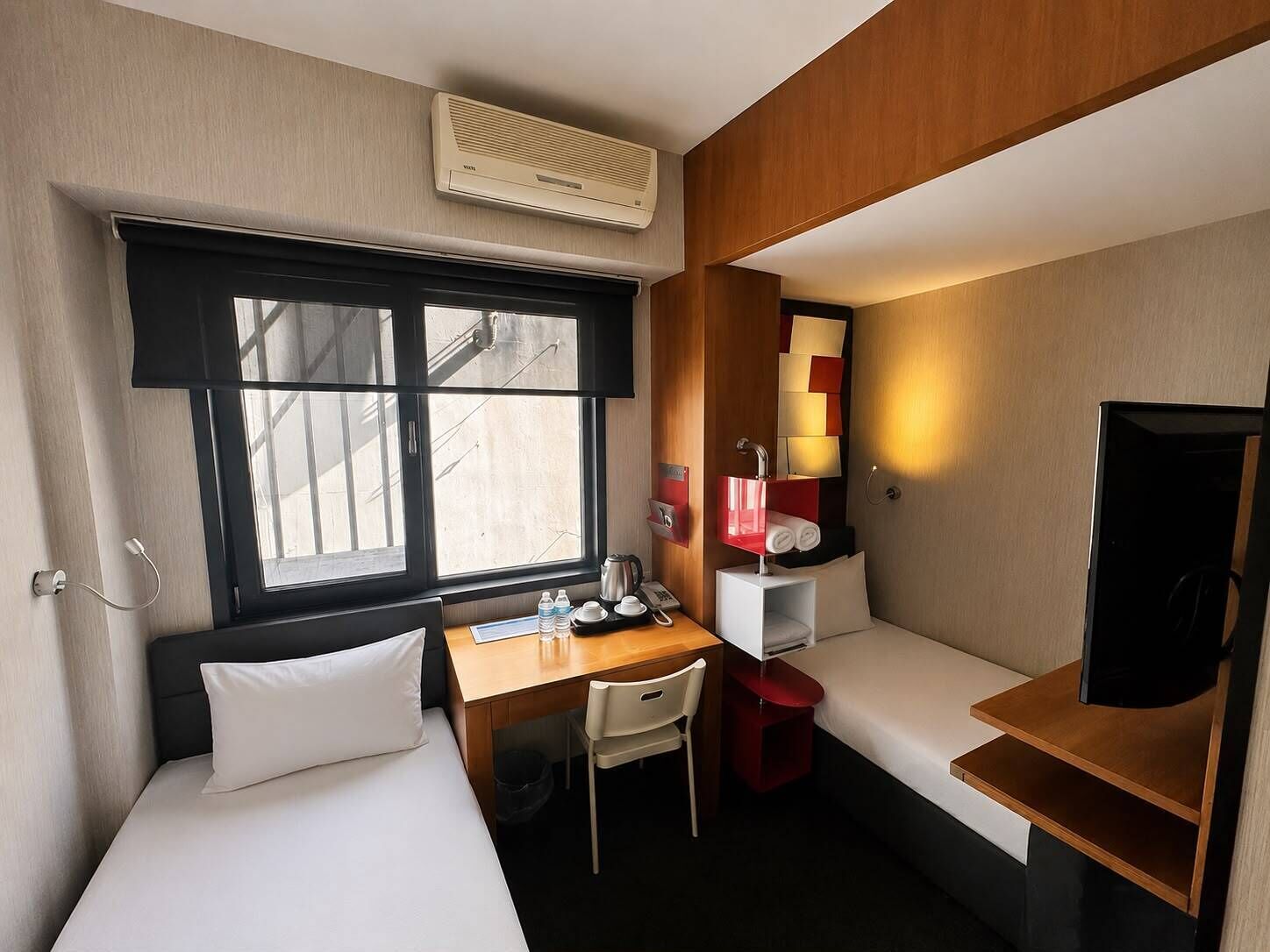 A compact hotel room with a bed, a desk, a chair, a small table, a TV, a window, and a wall-mounted air conditioner.