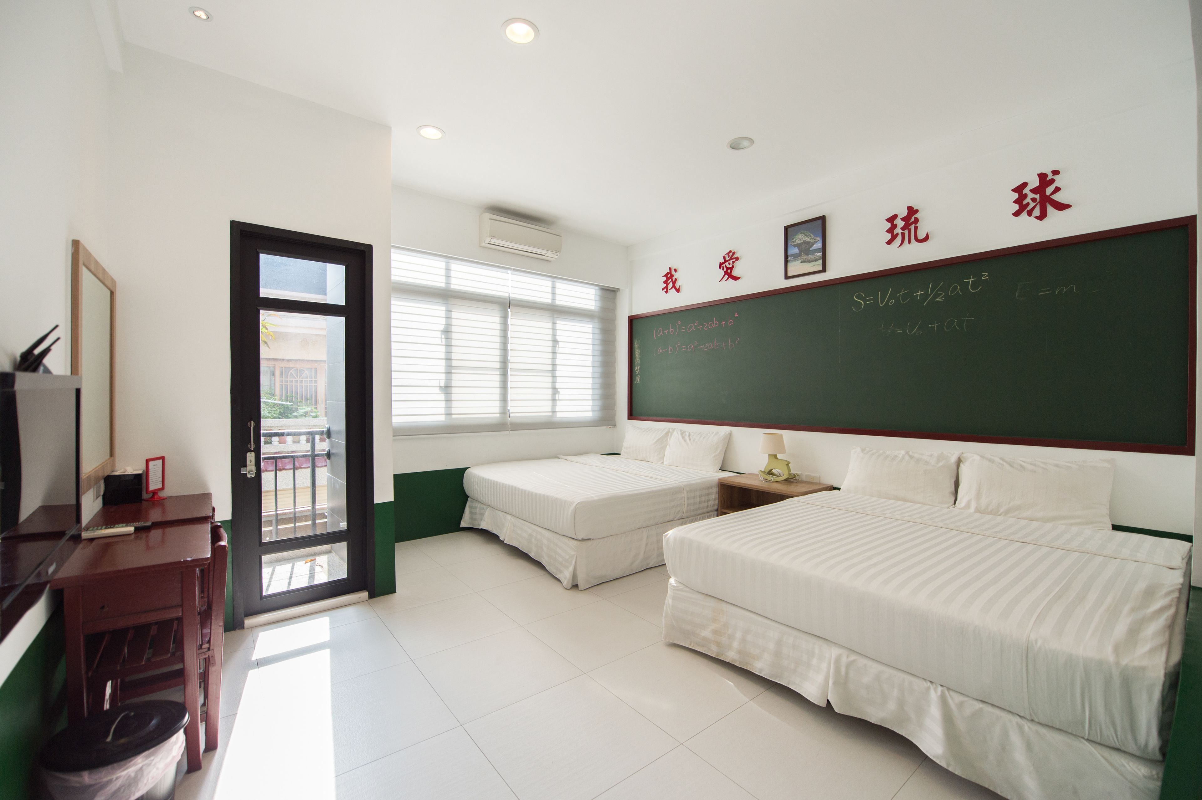 A classroom with a blackboard, two beds, a desk, and a door leading outside.