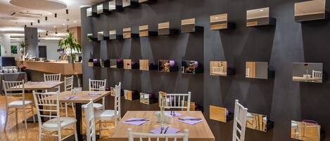 A modern restaurant interior with wooden tables and white chairs, a dark wall with mirrors and shelves, and a bar area in the background.
