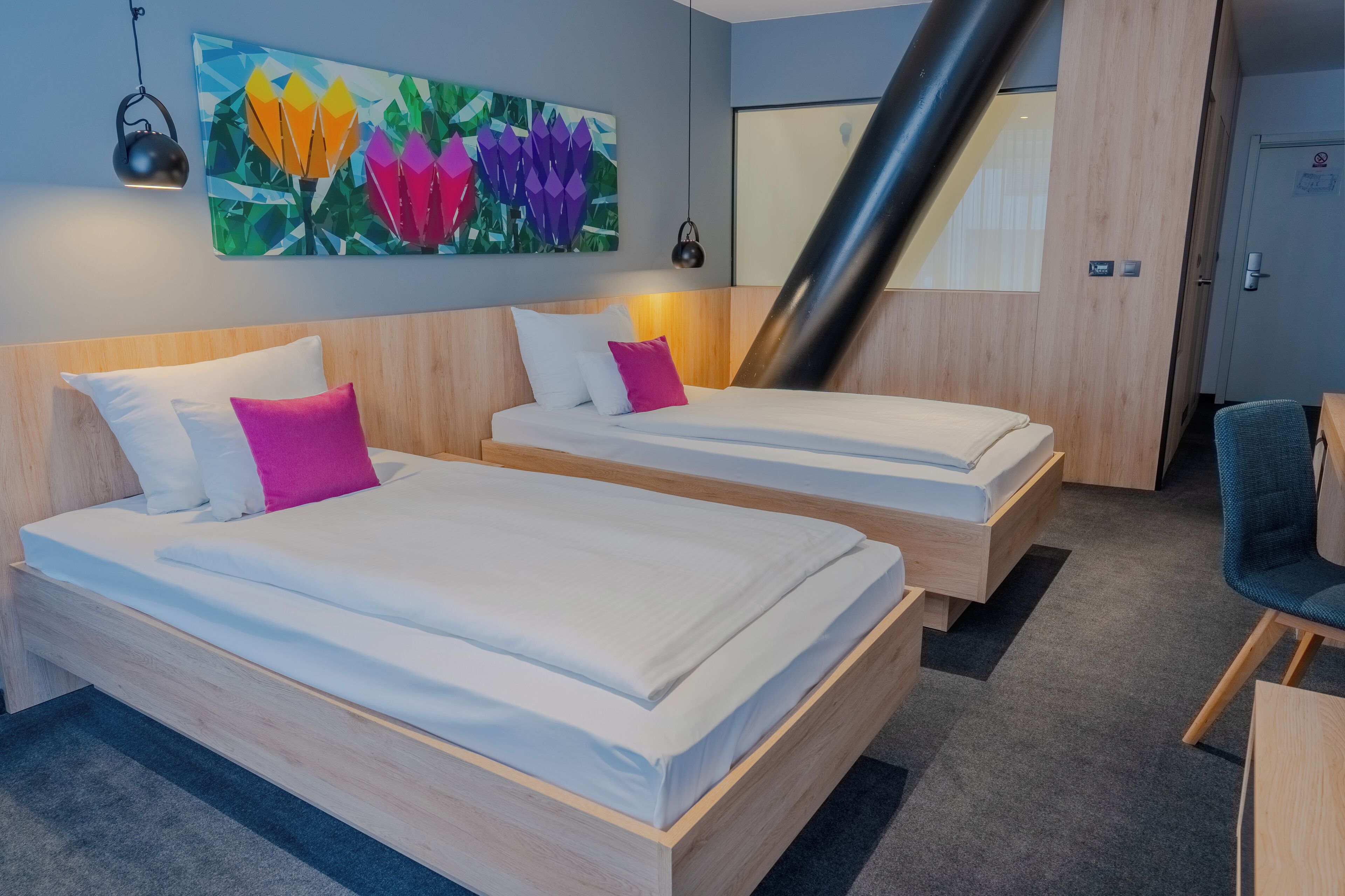 A hotel room with two beds, a wooden headboard, a colorful abstract painting, a black pipe, and a chair.