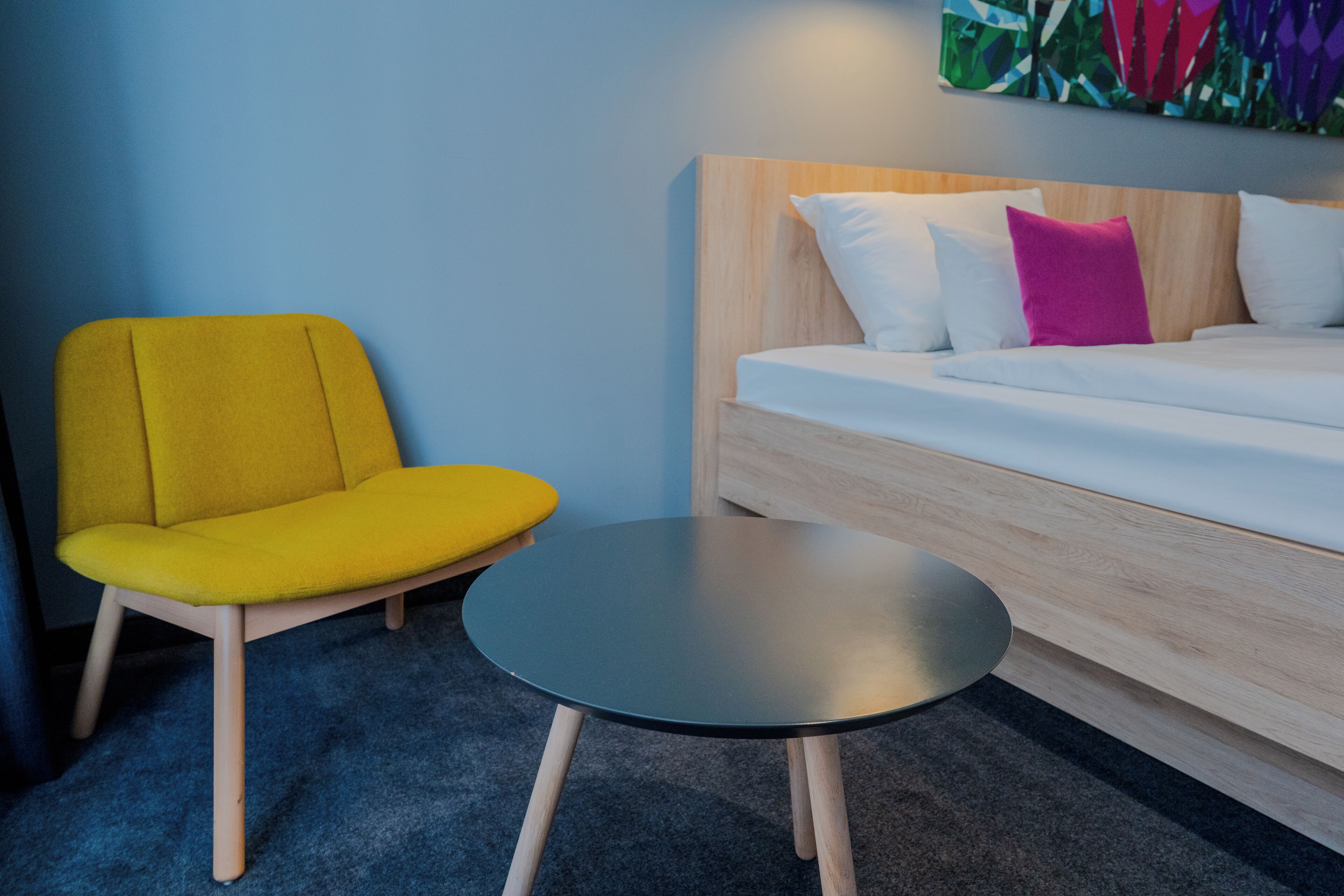 A modern hotel room with a bed, a yellow armchair, and a small round table.