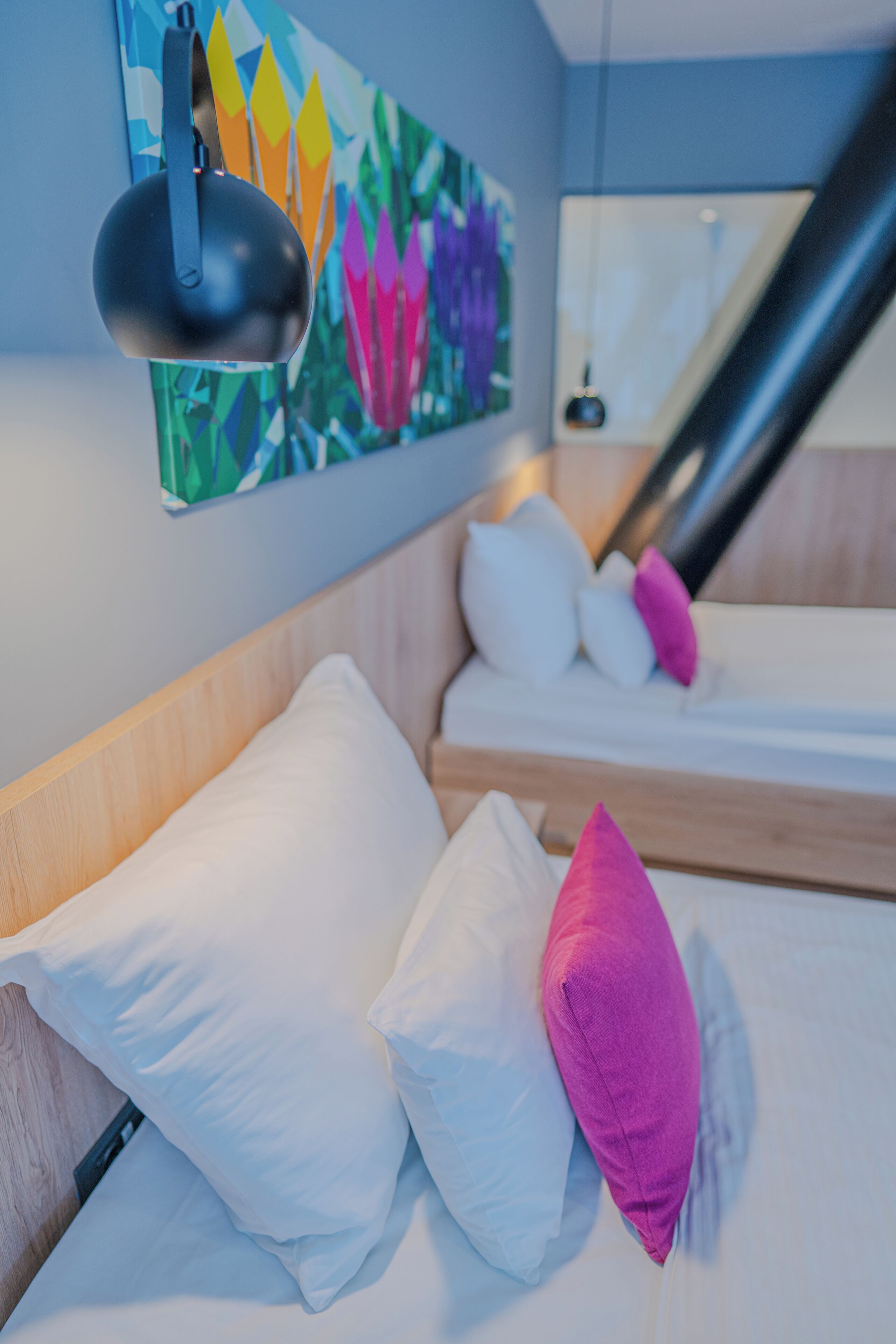 Habitación de hotel con cama, almohadas, una lámpara fijada a la pared y un cuadro de colores.