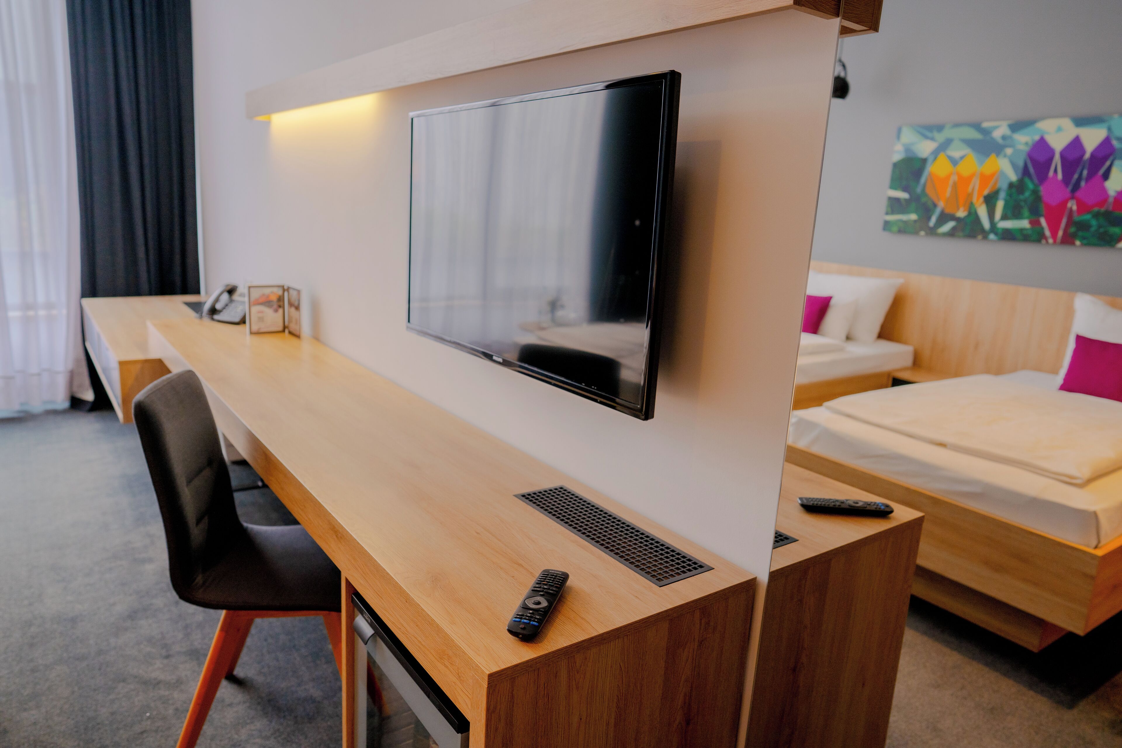 Ein Hotelzimmer mit einem Holztisch, einem an der Wand montierten Fernseher, einem Bett mit Kopfteil und einem farbenfrohen Kunstwerk an der Wand.