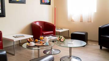 Una habitación de hotel moderna con un sillón de cuero rojo, una mesa de vidrio con desayuno y obras de arte en las paredes.