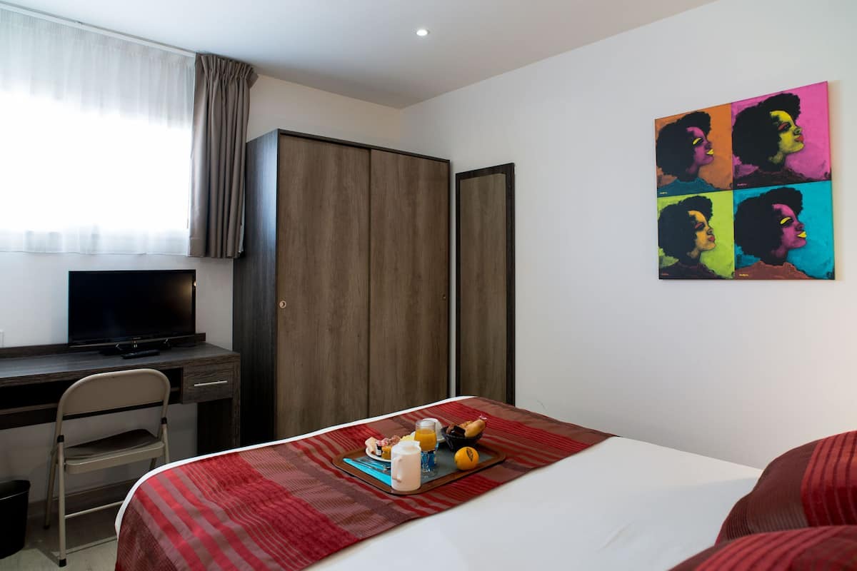 A hotel room with a bed, a desk with a TV, a wardrobe, and a colorful artwork on the wall.