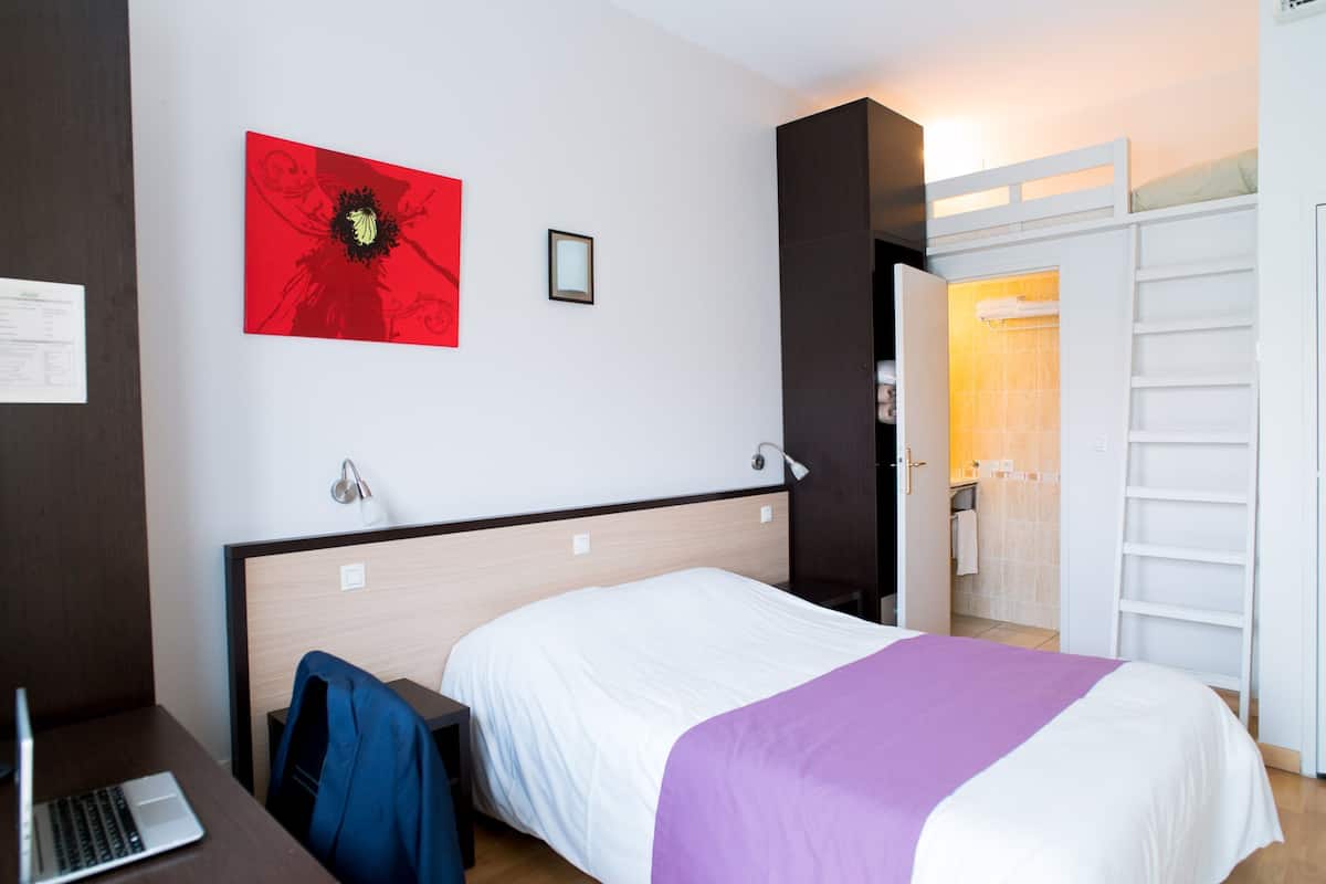 A hotel room with a bed, a desk with a laptop, a red artwork on the wall, and a bathroom visible through an open door.
