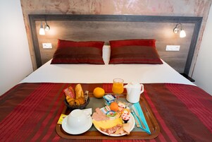 Una cama bien tendida con almohadas rojas y marrones, una bandeja de desayuno con pan, fruta, huevos y una jarra de jugo de naranja.