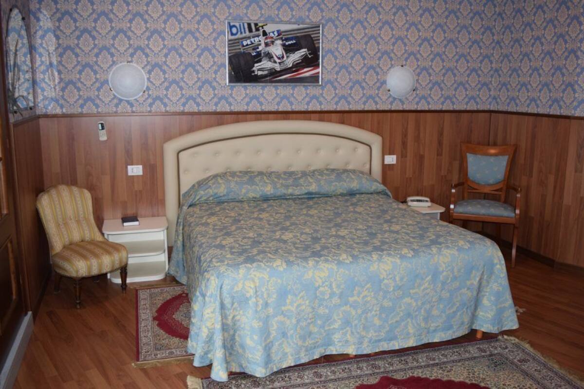 Une chambre à coucher avec un lit, deux chaises, une table de chevet, une horloge murale et une affiche.