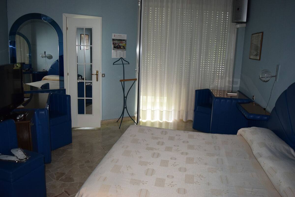 Une chambre d’hôtel comprenant un lit, deux fauteuils bleus, un bureau avec une chaise, un miroir, une fenêtre avec des rideaux et un climatiseur fixé au mur.