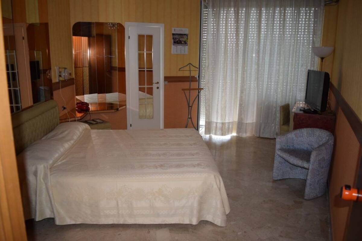 Une chambre à coucher avec un grand lit, une télévision, une chaise, un miroir et une porte donnant sur une autre pièce.