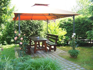 A wooden bench and table under a canopy in a garden with a brick pathway.