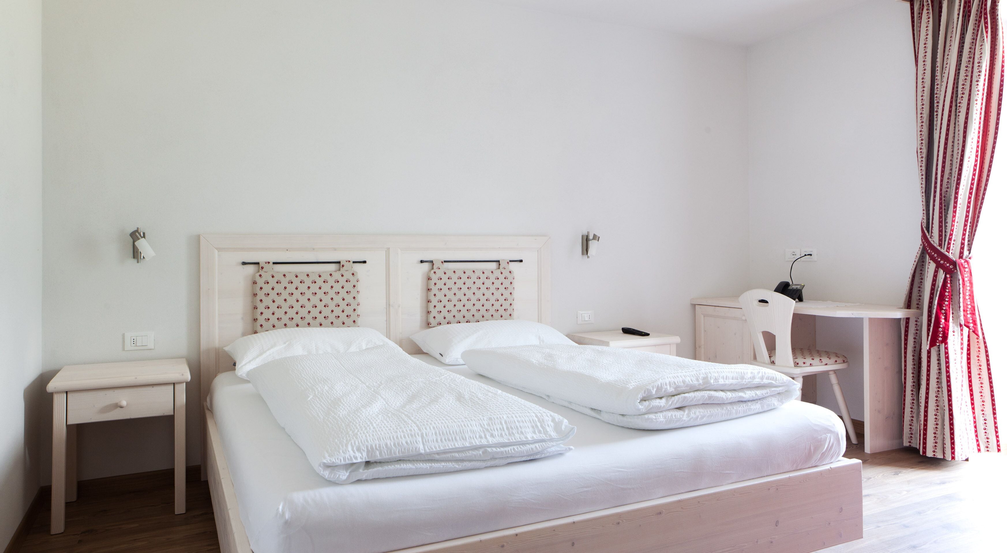Una cama bien tendida con sábanas blancas, una mesita de noche con una lámpara, una silla y un escritorio con un ordenador.