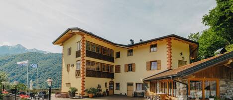 A two-story building with a courtyard, featuring a stone facade, wooden shutters, and a mountain backdrop.