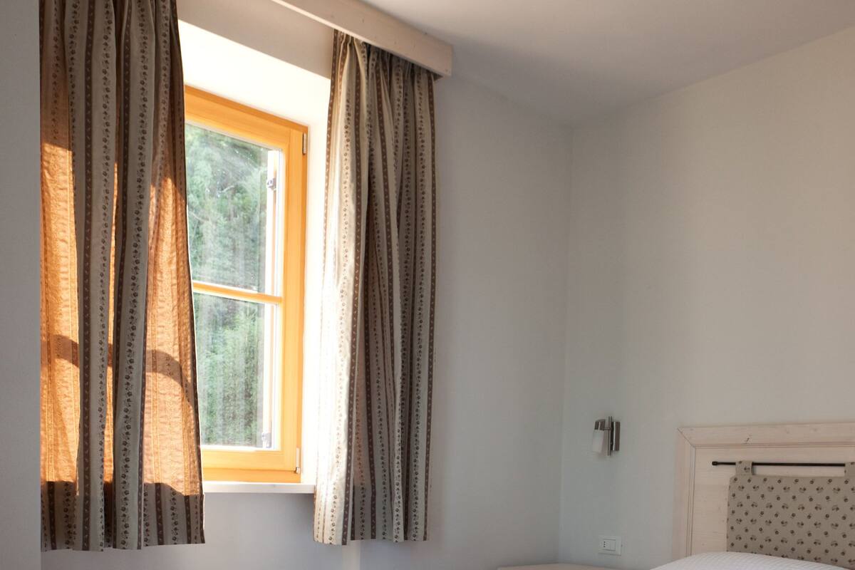 A bedroom with a wooden floor, a bed with white bedding, a window with curtains, and a small bedside table.