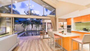 A modern kitchen with a large island, a dining area with chairs, and a view of a swimming pool and greenery. - 2 Bedrooms Villa Aperol (Koh Samui)