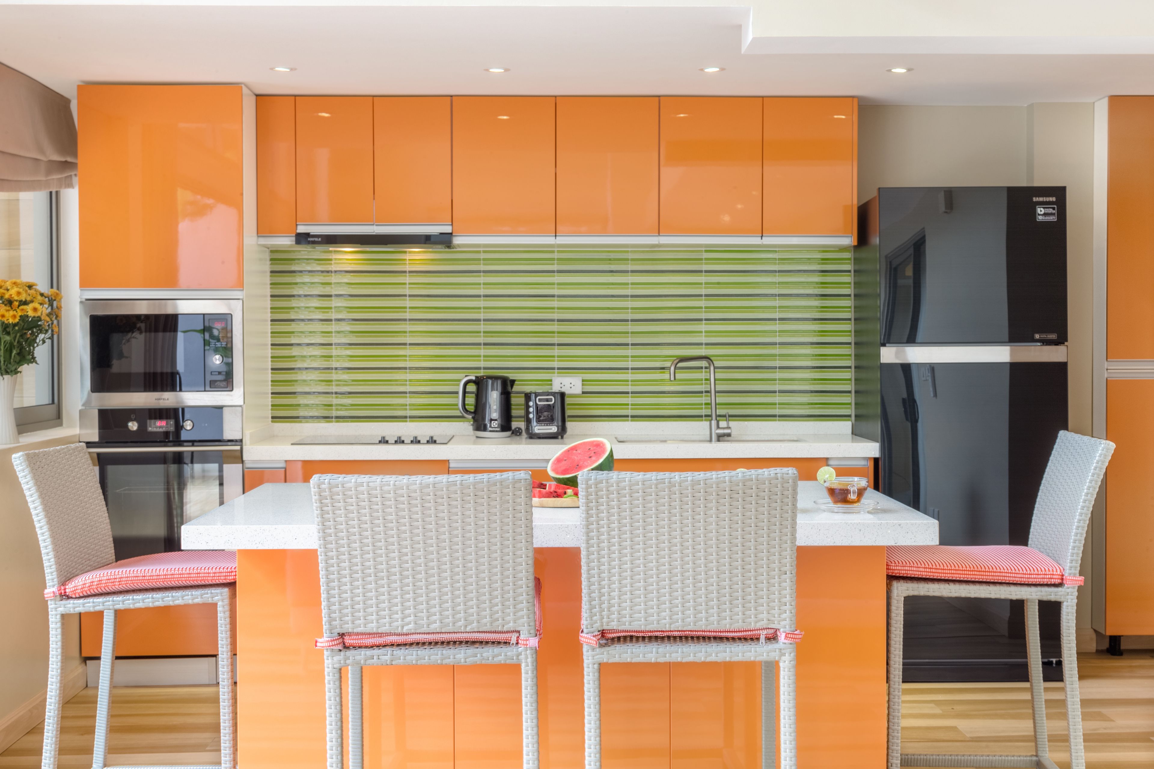 A modern kitchen with orange cabinets, a green-striped backsplash, a stainless steel refrigerator, and a dining area with wicker chairs.