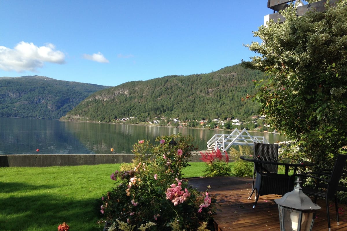 A lakeside deck with a railing, a table, and chairs, overlooking a calm lake and forested hills.