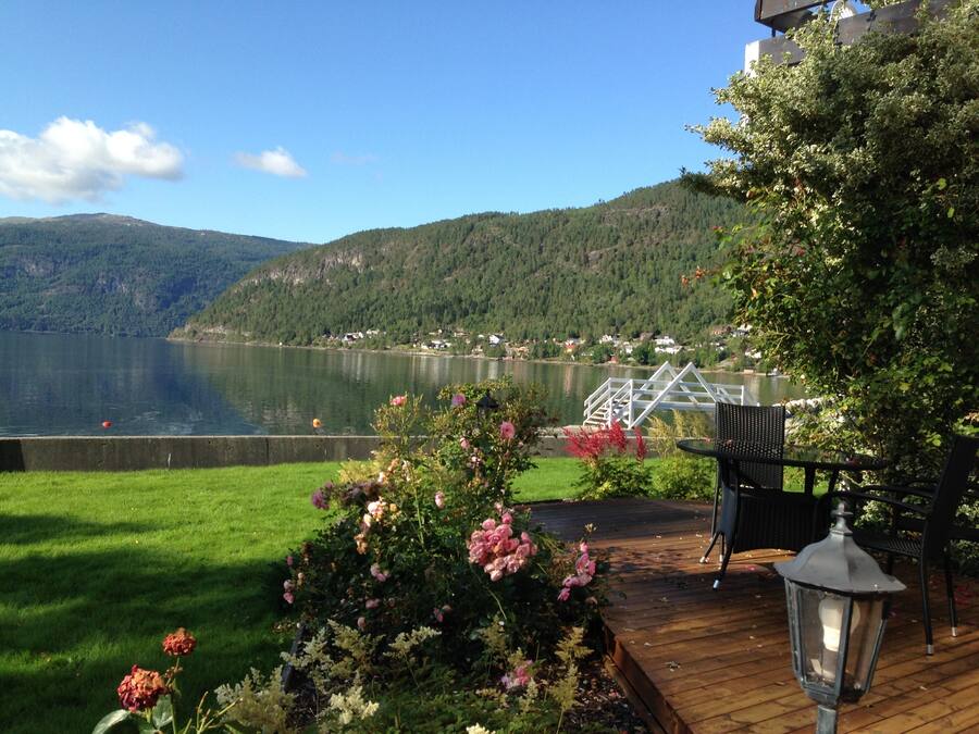 A lakeside deck with a railing, a table, and chairs, overlooking a calm lake and forested hills.