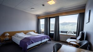Et hotelværelse med seng, skrivebord, stol og udsigt til sø og bjerge.