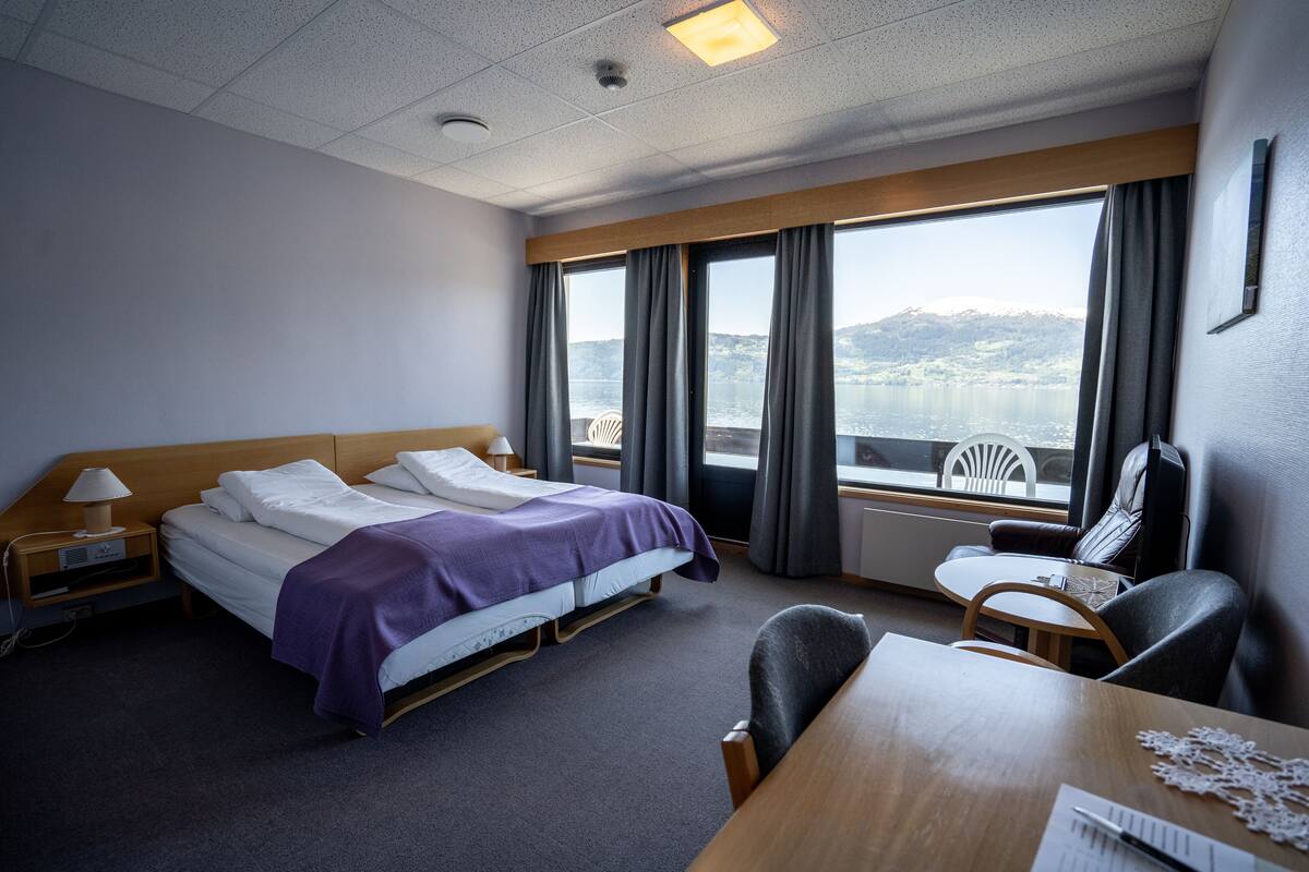 A hotel room with a bed, a desk, a chair, and a view of a lake and mountains.