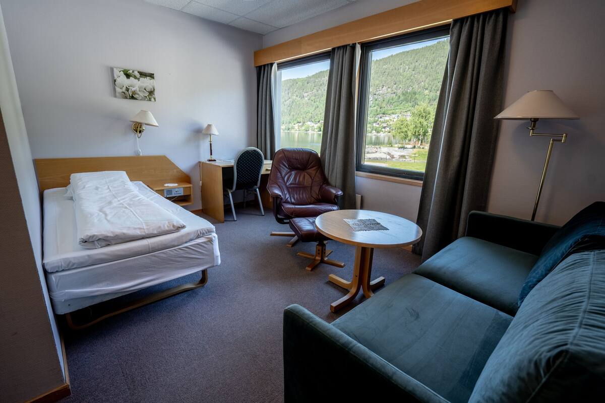 A hotel room with a bed, a sofa, a chair, a small table, and a window with a view of a lake and hills.
