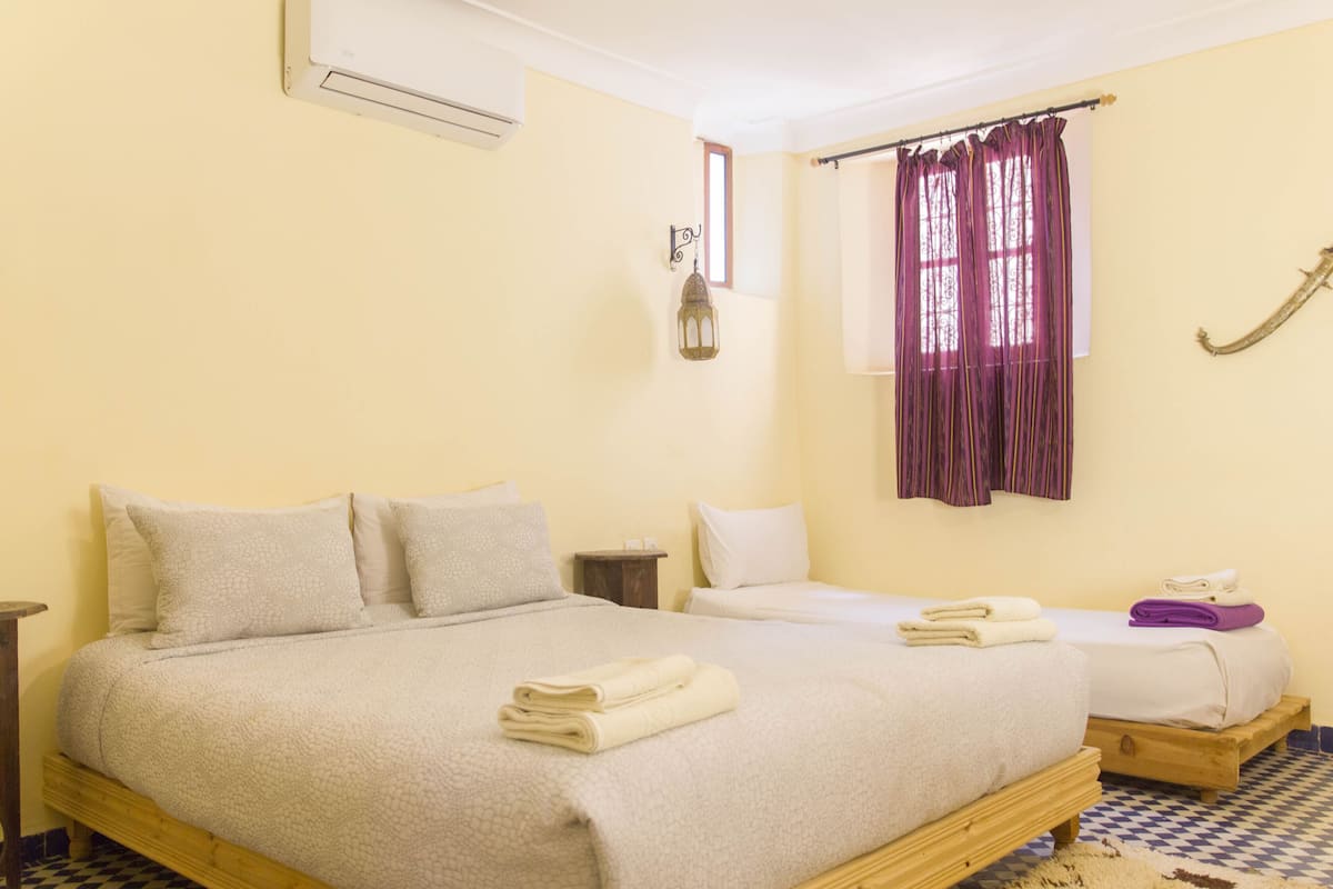 Una habitación de hotel con una cama grande, una cama más pequeña, una mesita de noche, una ventana con cortinas y una lámpara de pared.