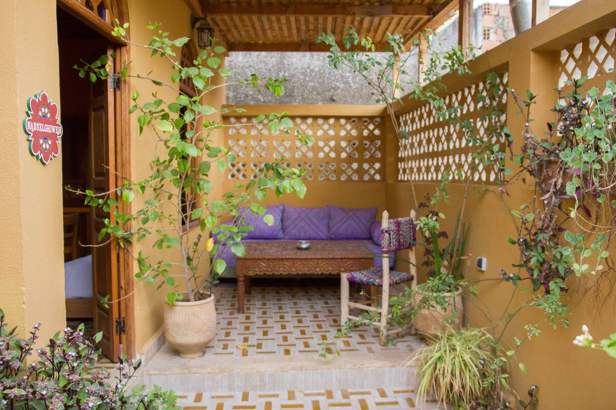 Un espacio al aire libre acogedor con un banco de madera y cojines, rodeado de plantas y un muro de celosía.