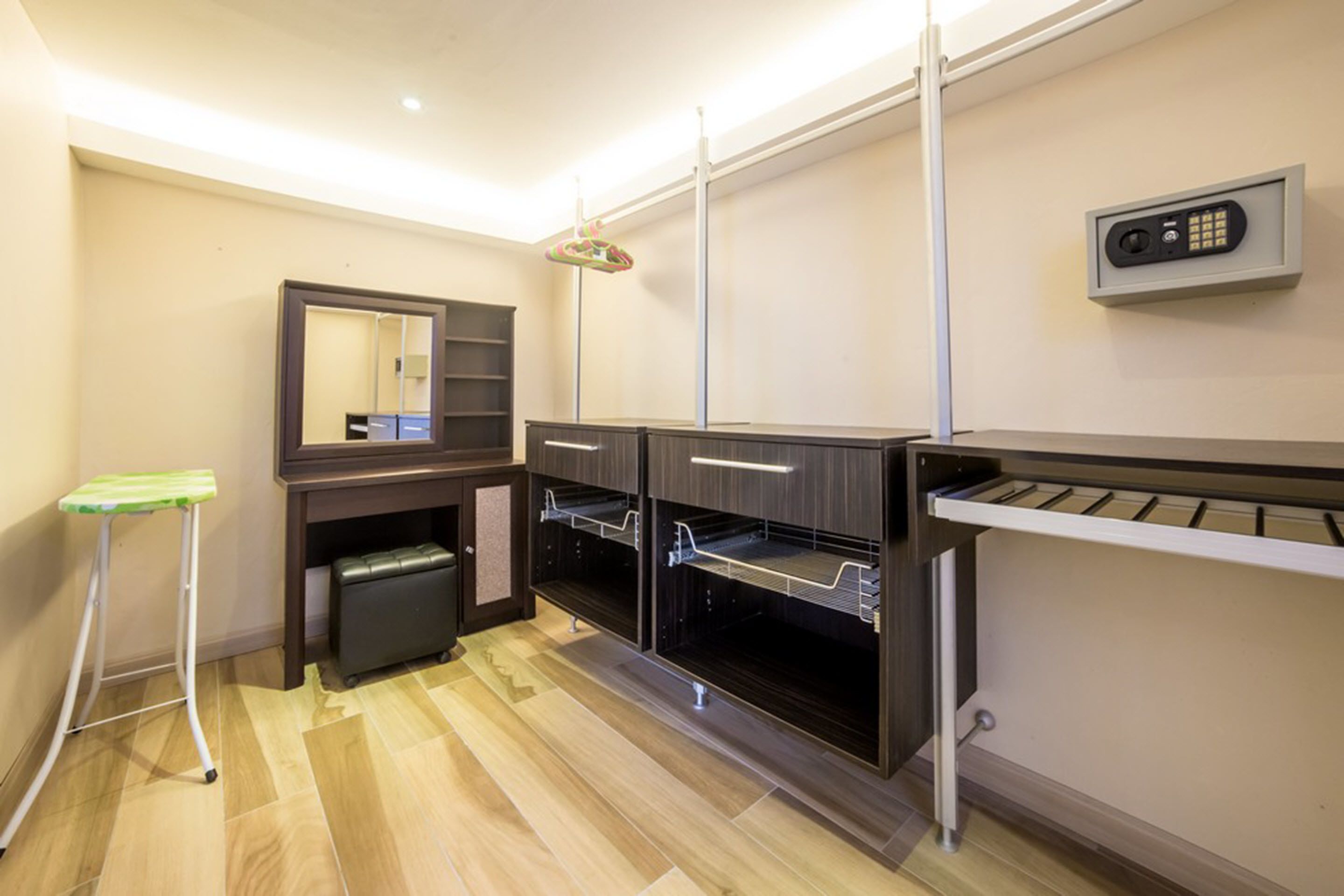 A hotel room with a wooden floor, a small stool, a mirror, a shelf unit, a hanging light fixture, and a wall-mounted security device.