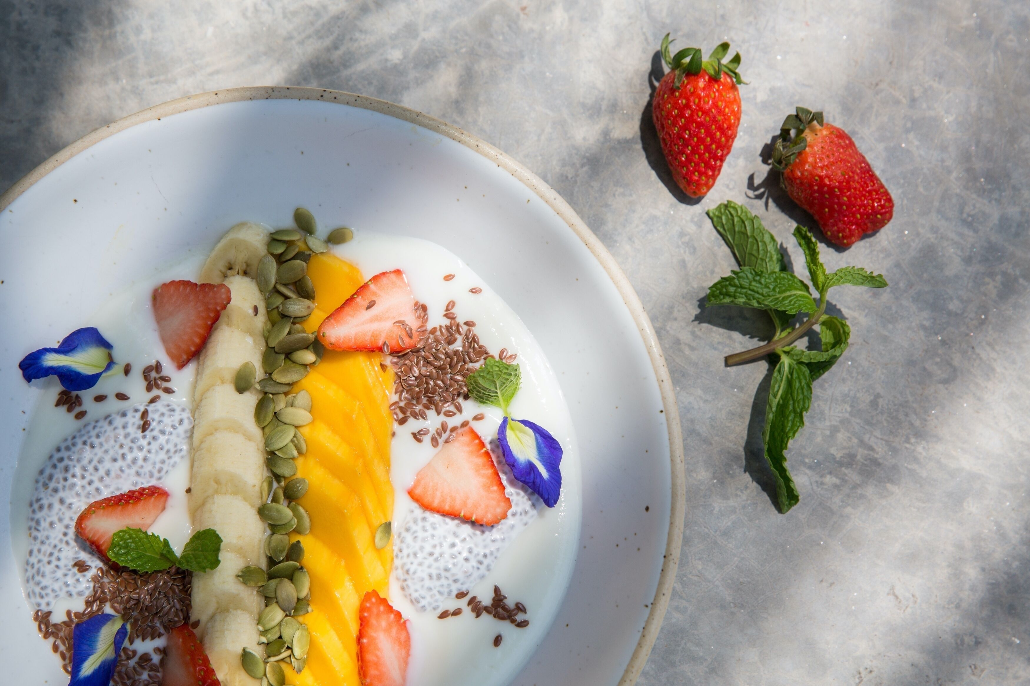 Een fruitsalade met aardbeien, bananen, mango en zaden, met een muntblaadje op een bord.