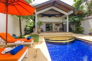 Uma área de piscina com espreguiçadeiras e um guarda-sol, um alpendre coberto com uma mesa de jantar e uma cozinha.
