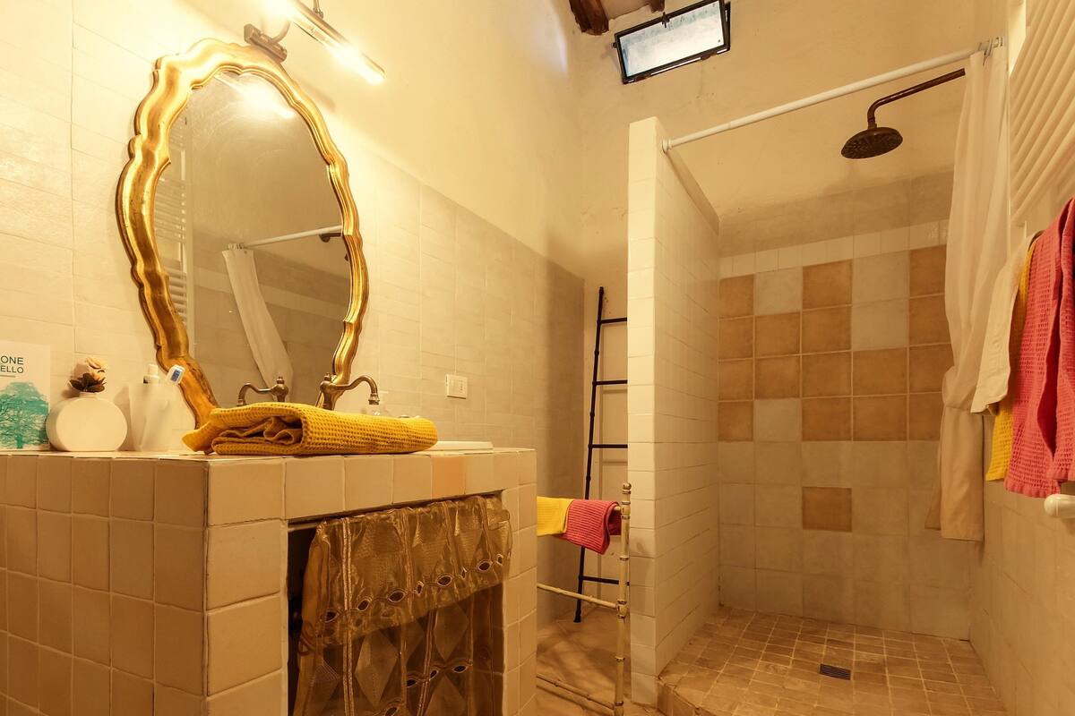 Ein Badezimmer mit gefliestem Boden, einem Waschbecken mit goldfarbenem Spiegelrahmen, einer Duschkabine mit gefliester Wand und einem Handtuchhalter.