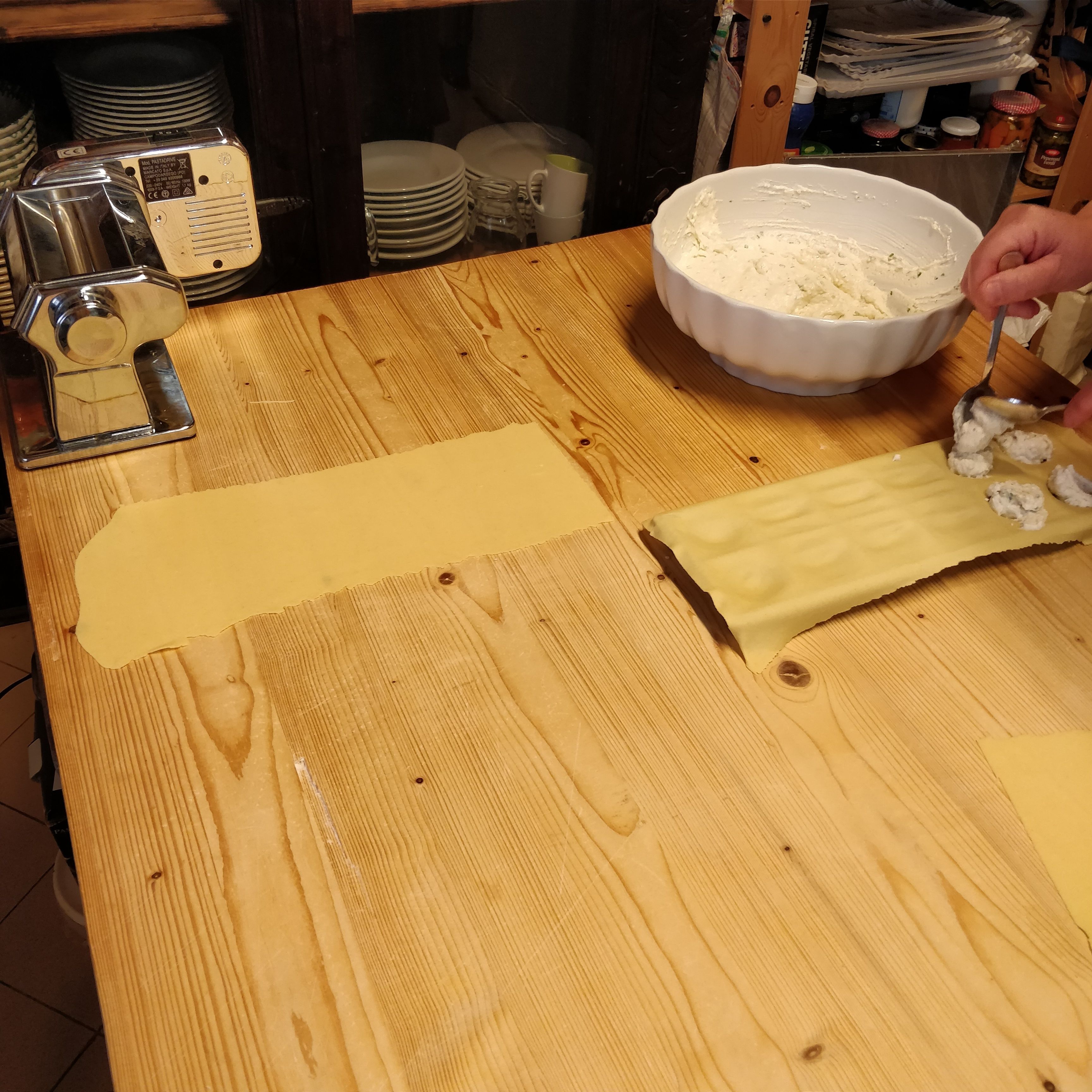 A kitchen counter with a pasta machine, a bowl of dough, and a person preparing dough.