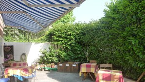An outdoor patio area with tables and chairs under a striped awning, surrounded by greenery. - Locanda Gaia (Muggia)