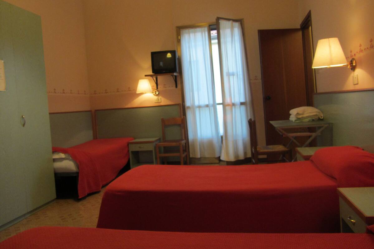 A hotel room with two single beds, a desk, a chair, a table, a lamp, and a television.