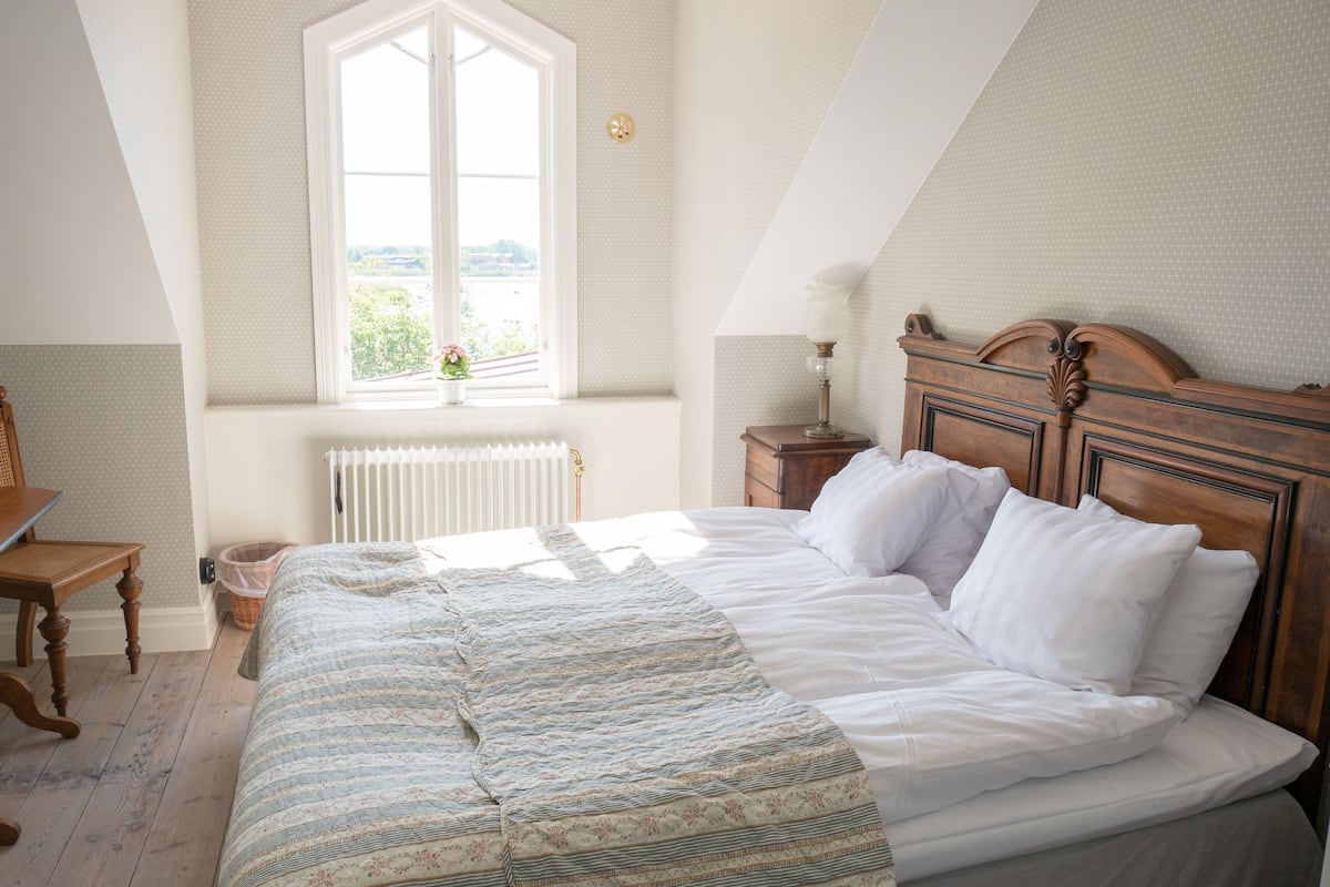 A bedroom with a large bed, a wooden headboard, a window with a view, a radiator, and a bedside table with a lamp.