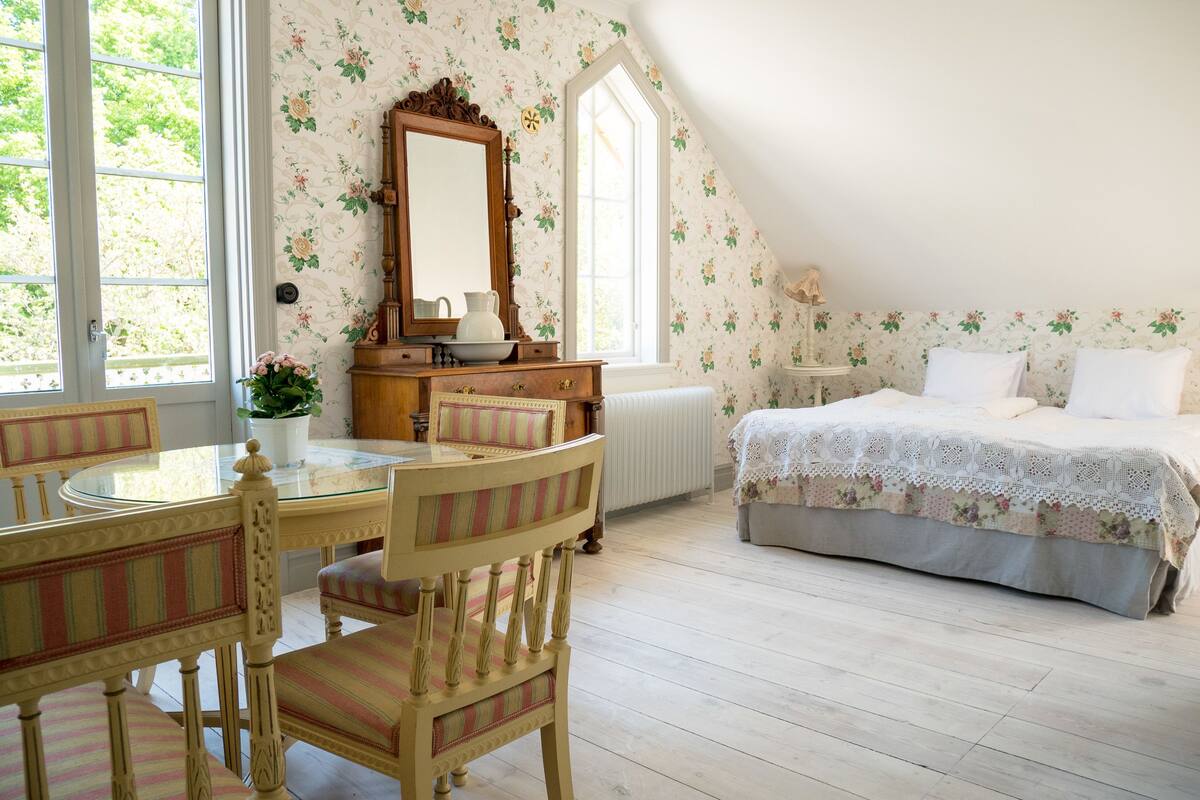 A bedroom with a bed, a dresser, a mirror, a window, and a dining area with chairs and a table.