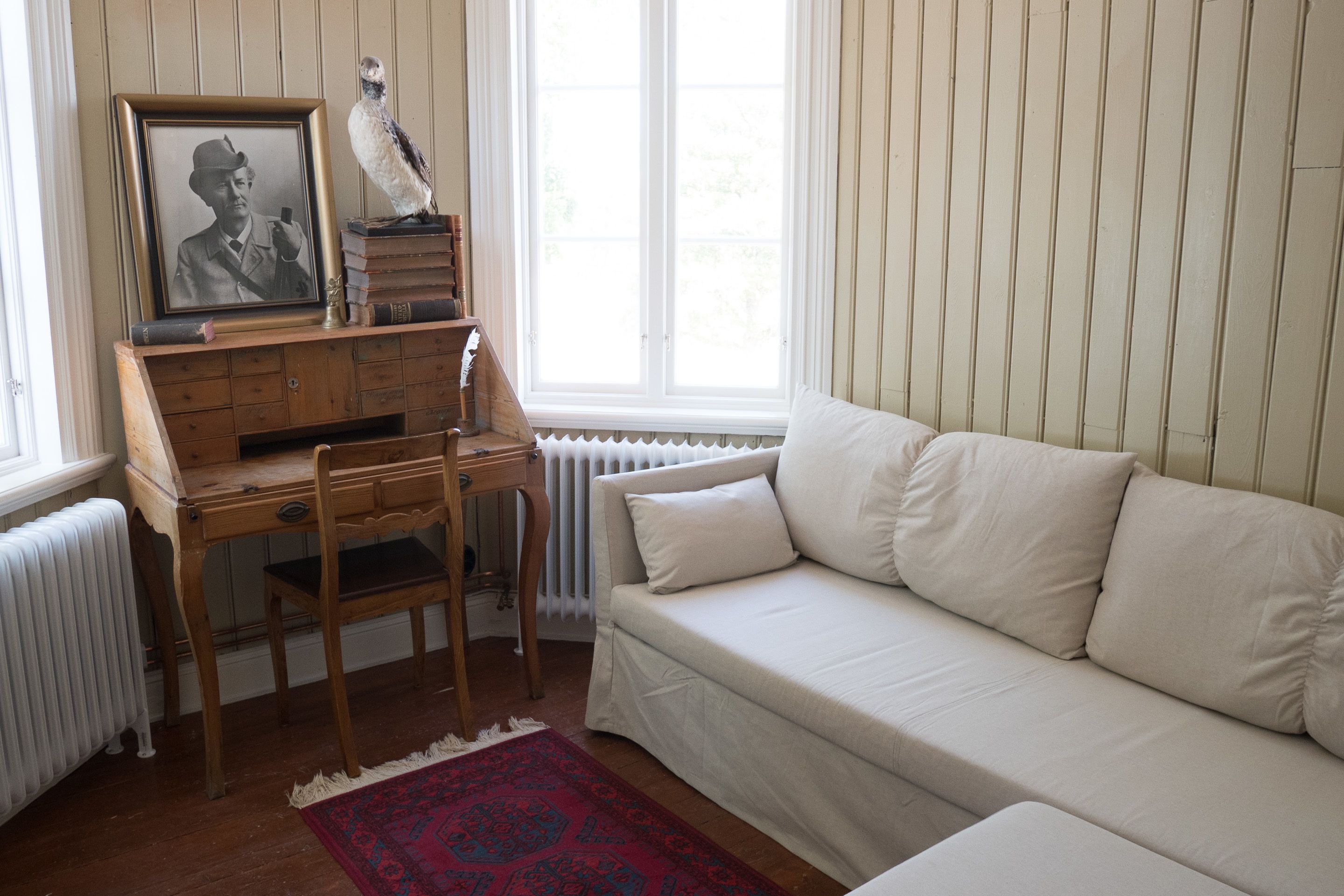 Una habitación con un escritorio de madera, un sofá blanco, una fotografía enmarco y una estatua de pájaro.