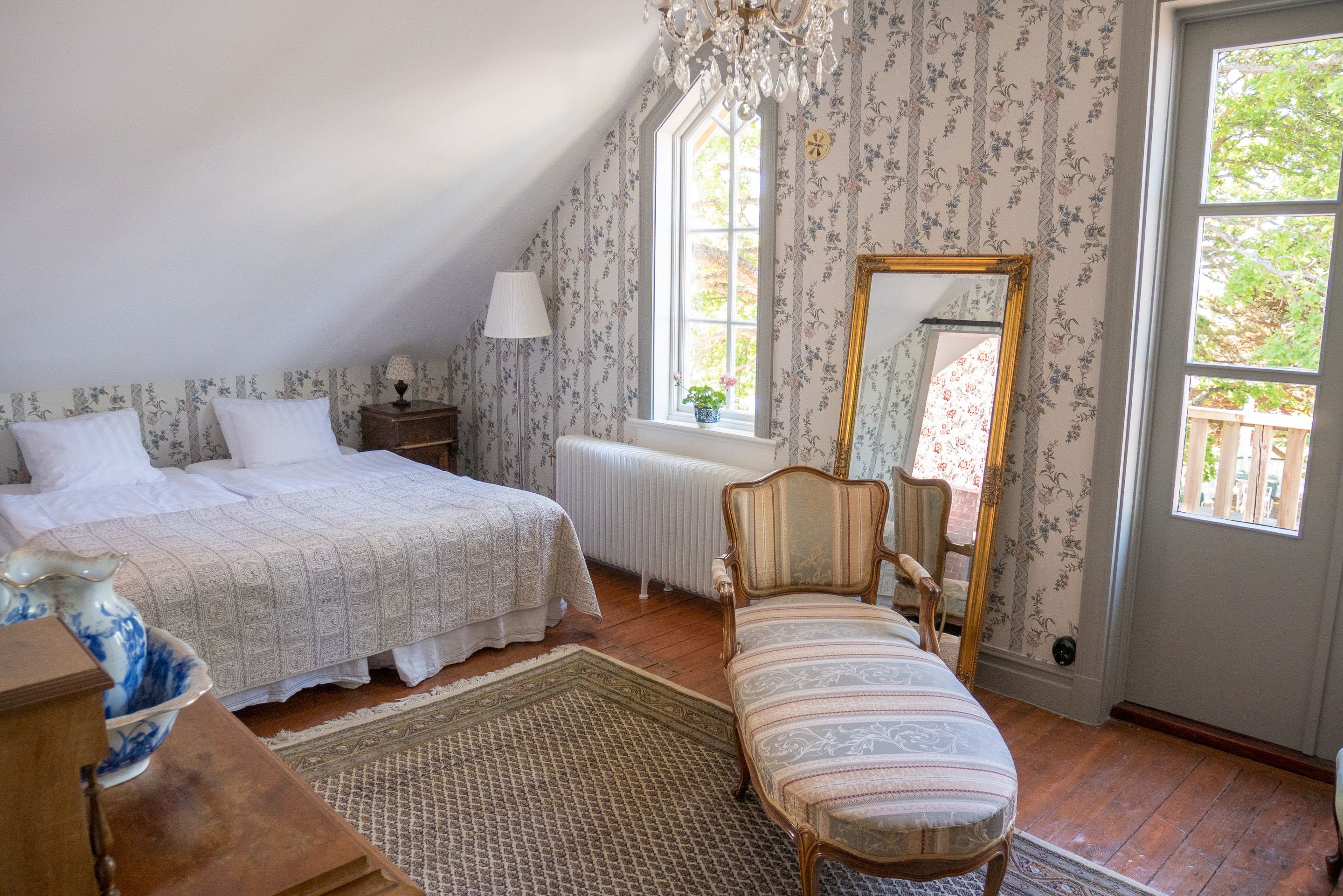 A bedroom with a bed, a chair, a dresser, a mirror, and a window with a view of greenery.