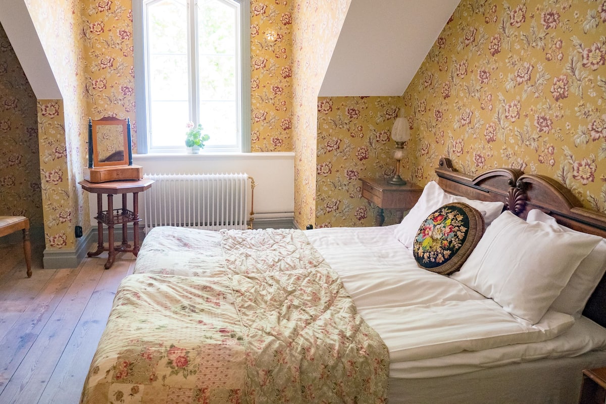 A bedroom with a bed, a small table, a chair, and a window with floral wallpaper.