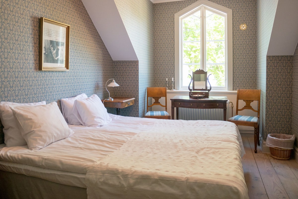 A bedroom with a large bed, a desk, two chairs, a window with a view, and a framed picture on the wall.