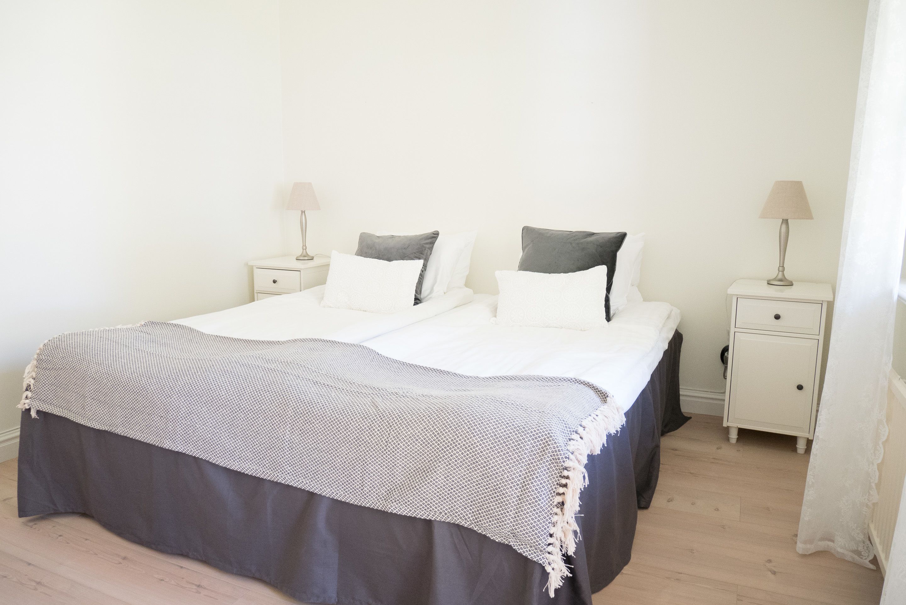 Una cama bien hecha con colcha gris y blanca, dos mesitas de noche con lámparas y un tocador blanco.