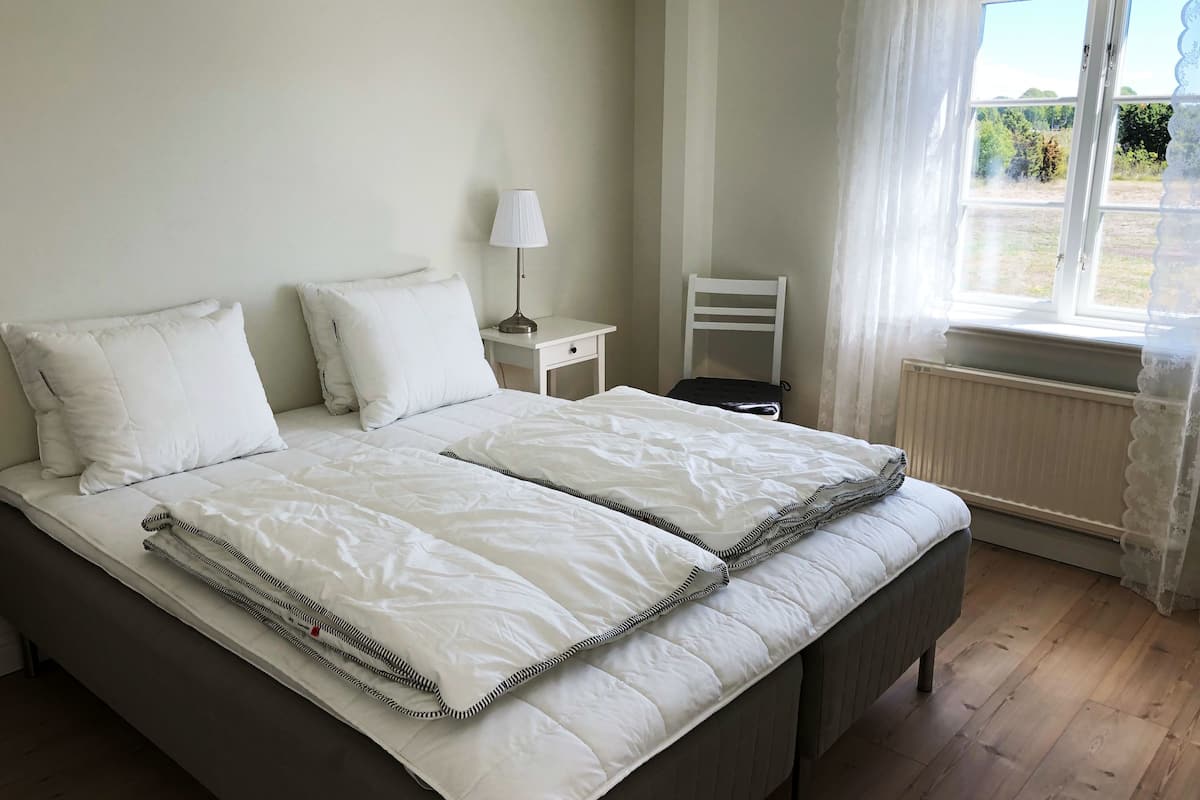 A double bed with white bedding, a bedside table with a lamp, a chair, and a window with curtains.