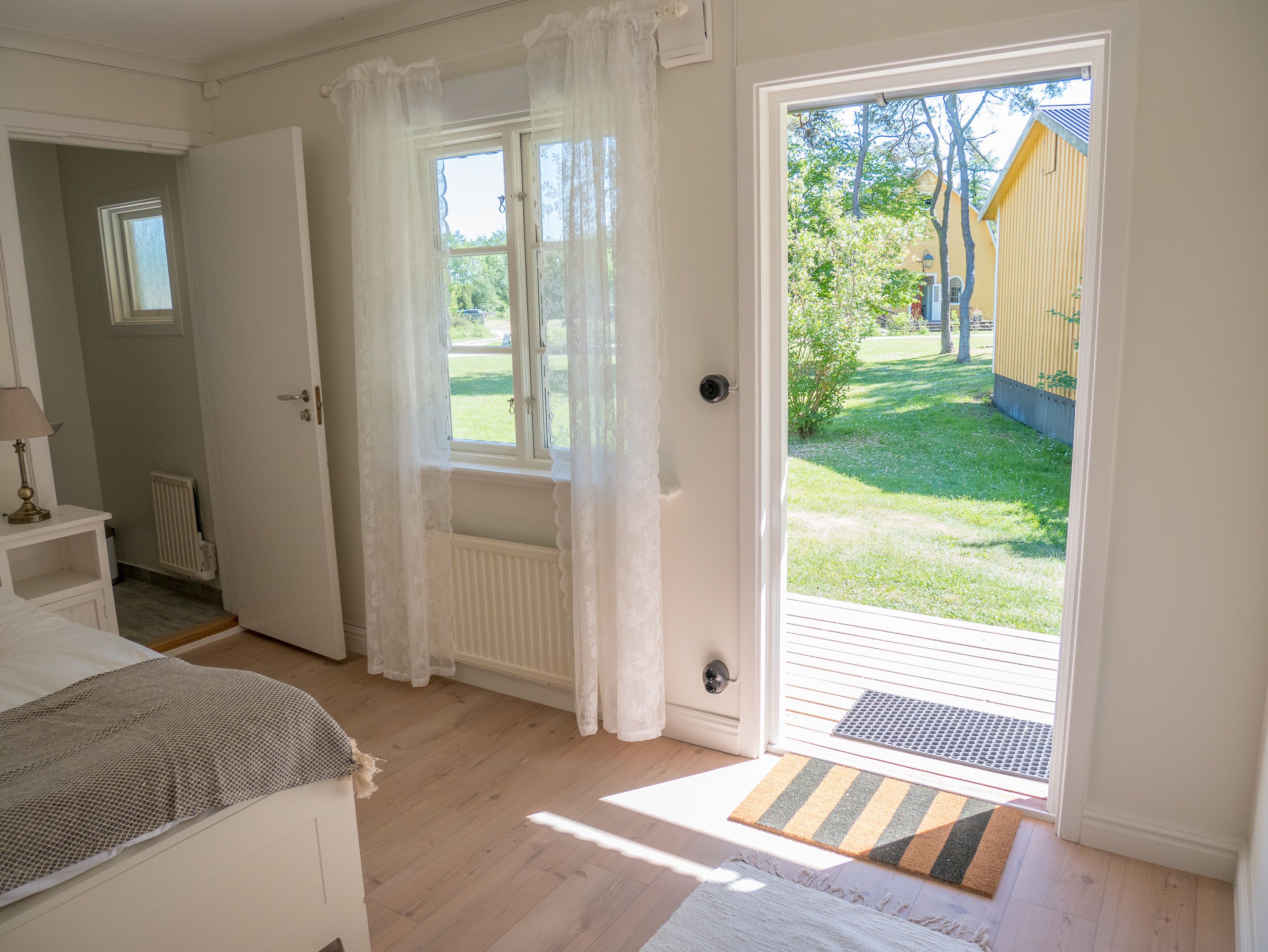 Een kamer met een bed, een raam met gordijnen, een radiator en uitzicht op een tuin en een geel gebouw.
