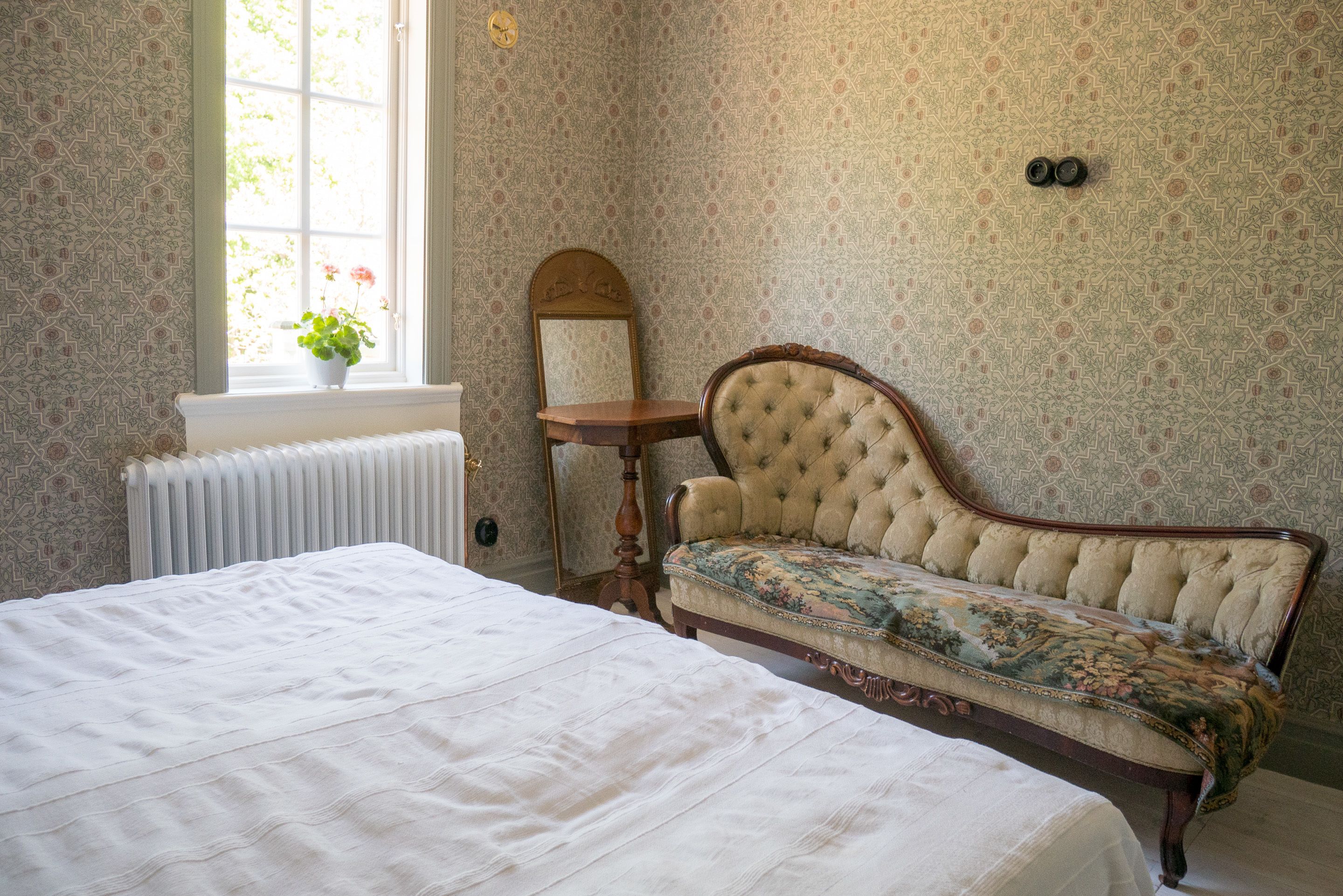 A bedroom with a bed, a window with a view, a radiator, a mirror, and a vintage sofa.