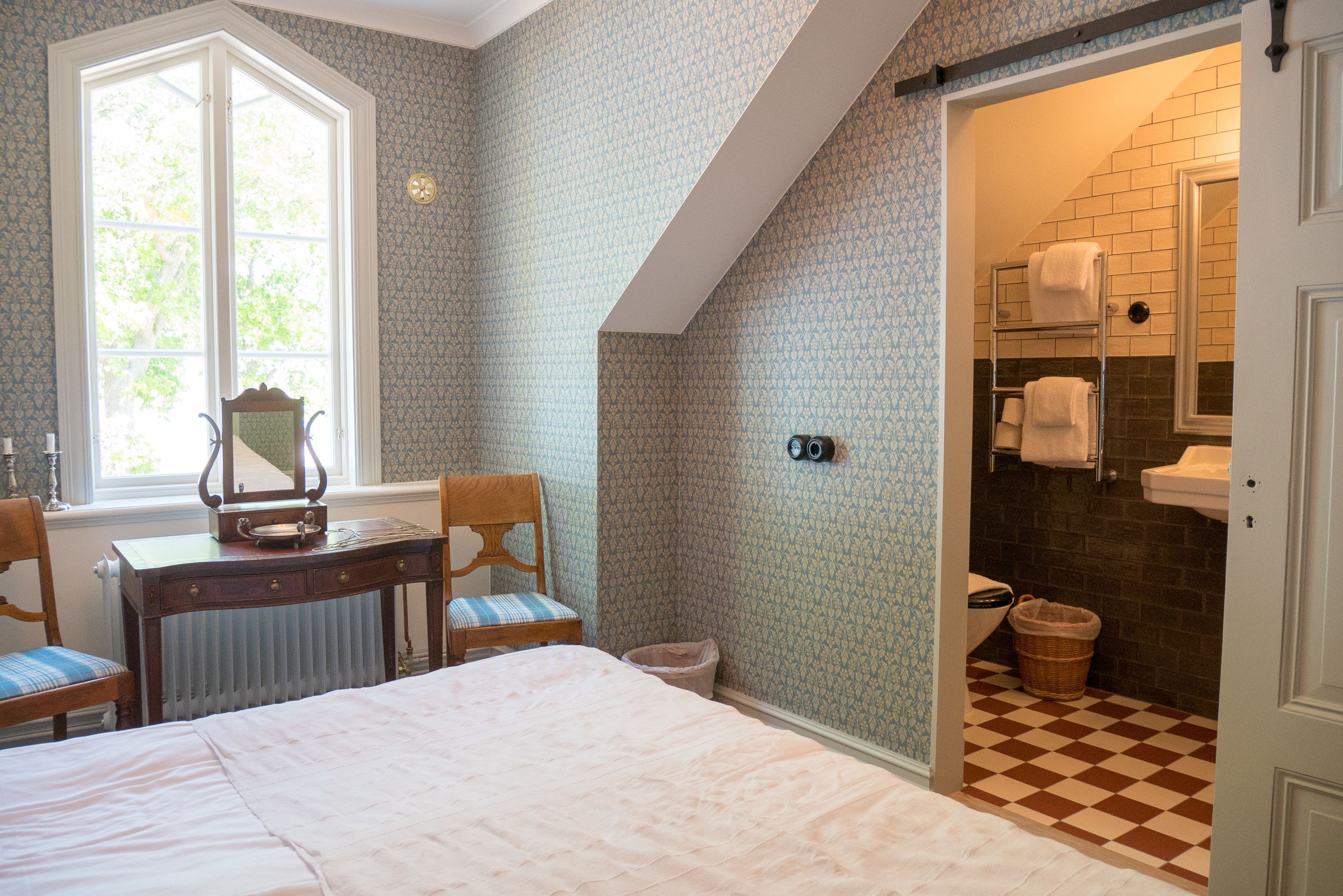 Un dormitorio con un ventanal grande, un escritorio con silla, una cama y un baño con piso a cuadros.