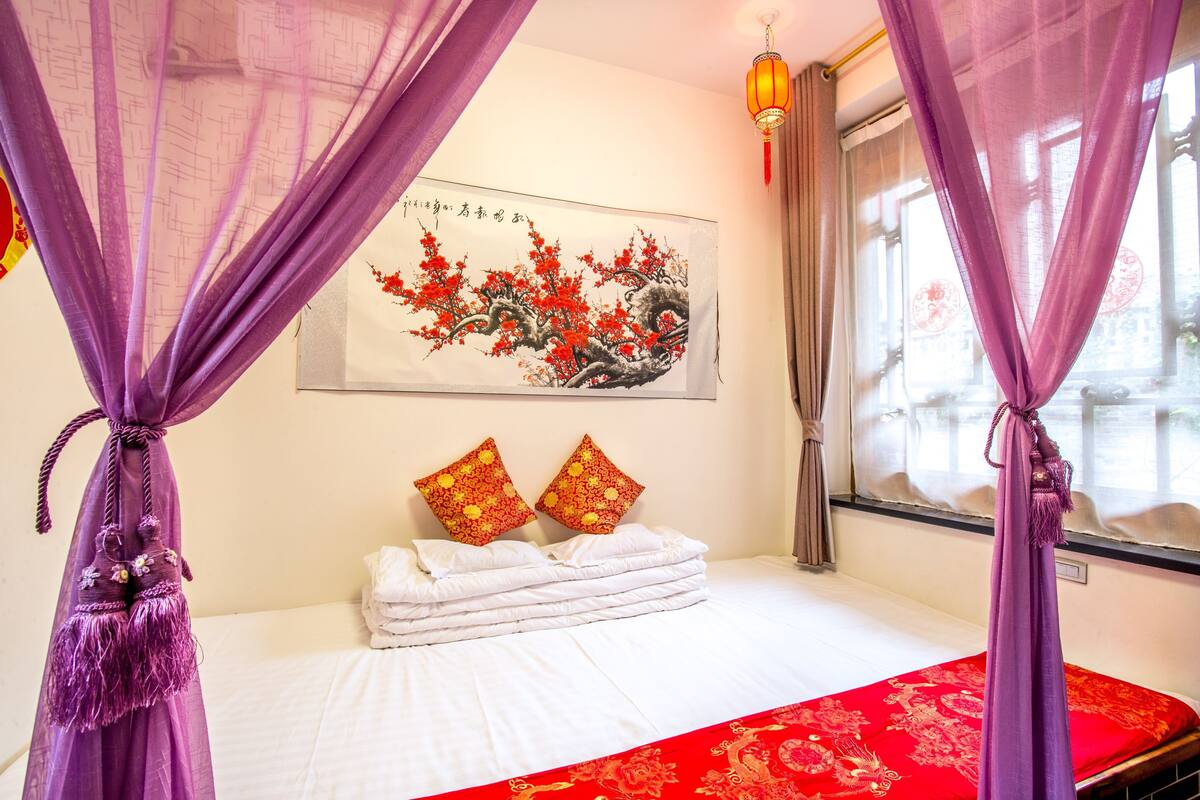 A hotel room with a bed, a red and white bedspread, a painting on the wall, and purple curtains.