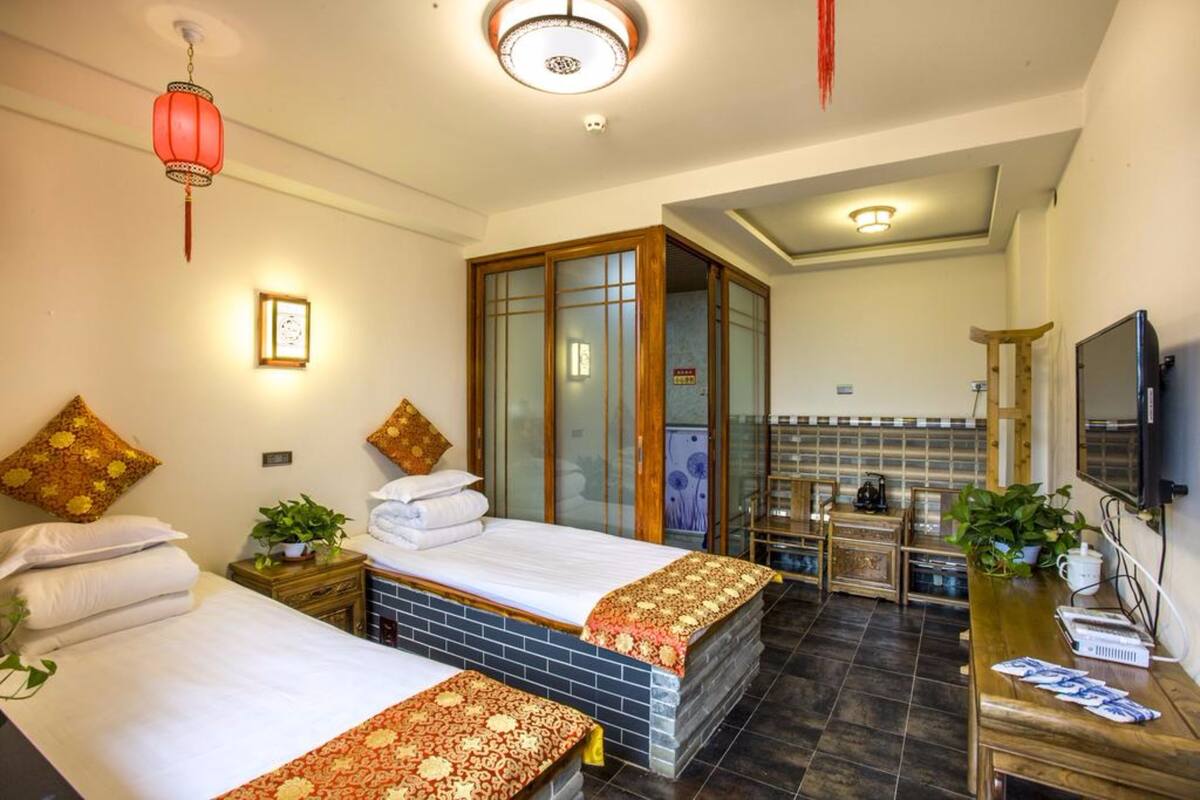 A hotel room with two beds, a wooden wardrobe, a TV, a desk with a plant, and a bathroom visible through a glass door.