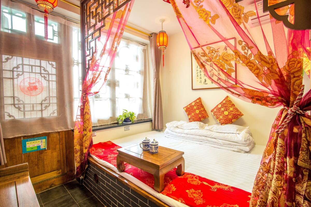 A traditional room with a bed, a small table with teapots, a window with curtains, and a wall with a framed artwork.